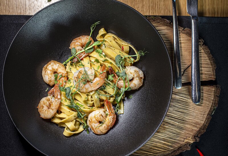 Tagliatelle z krewetkami, ziołowymi kiełkami i papryczką chili, podane w czarnej misce na rustykalnej desce ze sztućcami