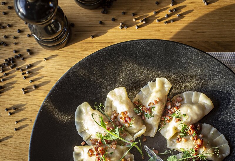 Pierogi w półksiężycowym kształcie na czarnym talerzu, z chrupiącymi skwarkami i kiełkami; widelec i nóż, młynki do pieprzu na drewnianym stole.