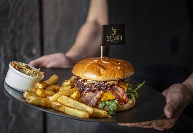 Burger z boczkiem i serem, świeże liście, frytki i colesław na czarnej tacy, mała flaga „Scandi”, podany przez obsługę