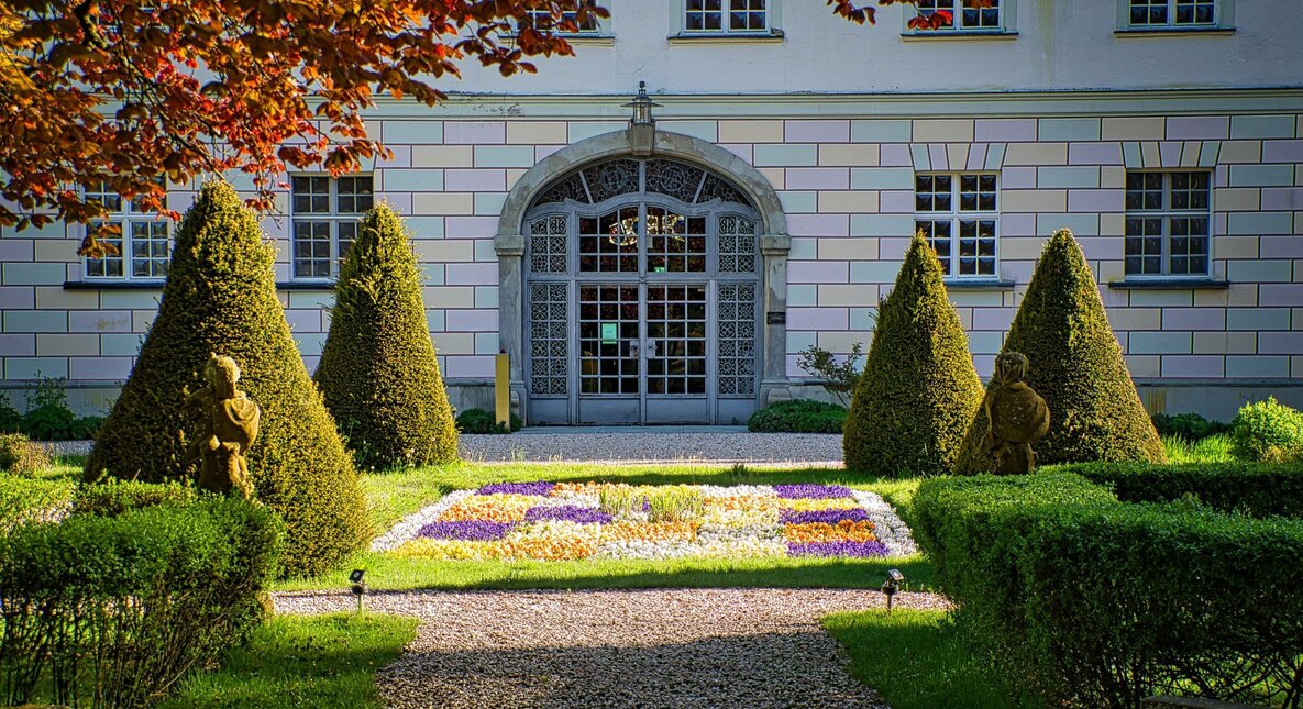 Manor garden with patterned flowerbed, cone-shaped topiary, gravel path and ornate arched glass entrance.