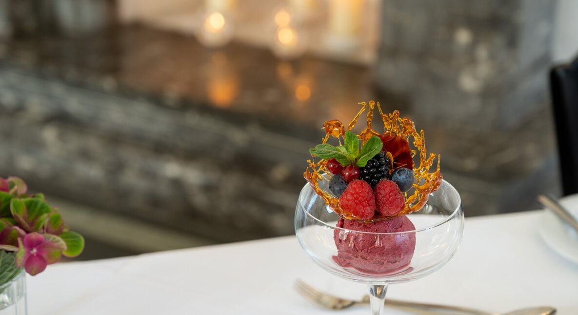 Berry sorbet in a stemmed glass topped with spun caramel, fresh berries, mint and edible flower on white tablecloth; candlelit fireplace.