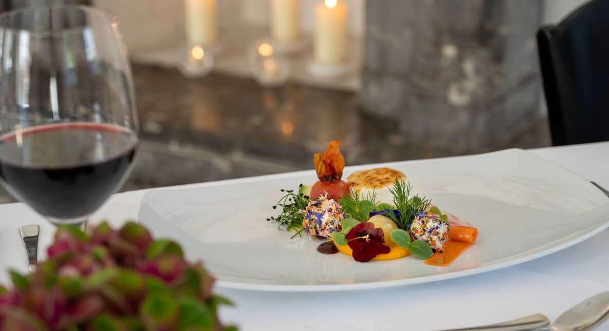 Fine-dining plated starter with edible flowers, microgreens and sauces on white plate, red wine glass and lit candles in background.
