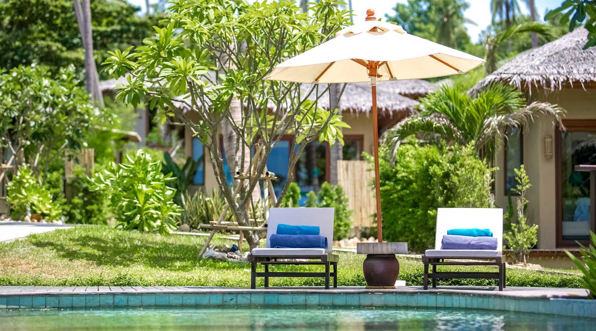 Poolside lounge chairs with towels and a parasol set in a lush tropical garden outside thatched-roof bungalows