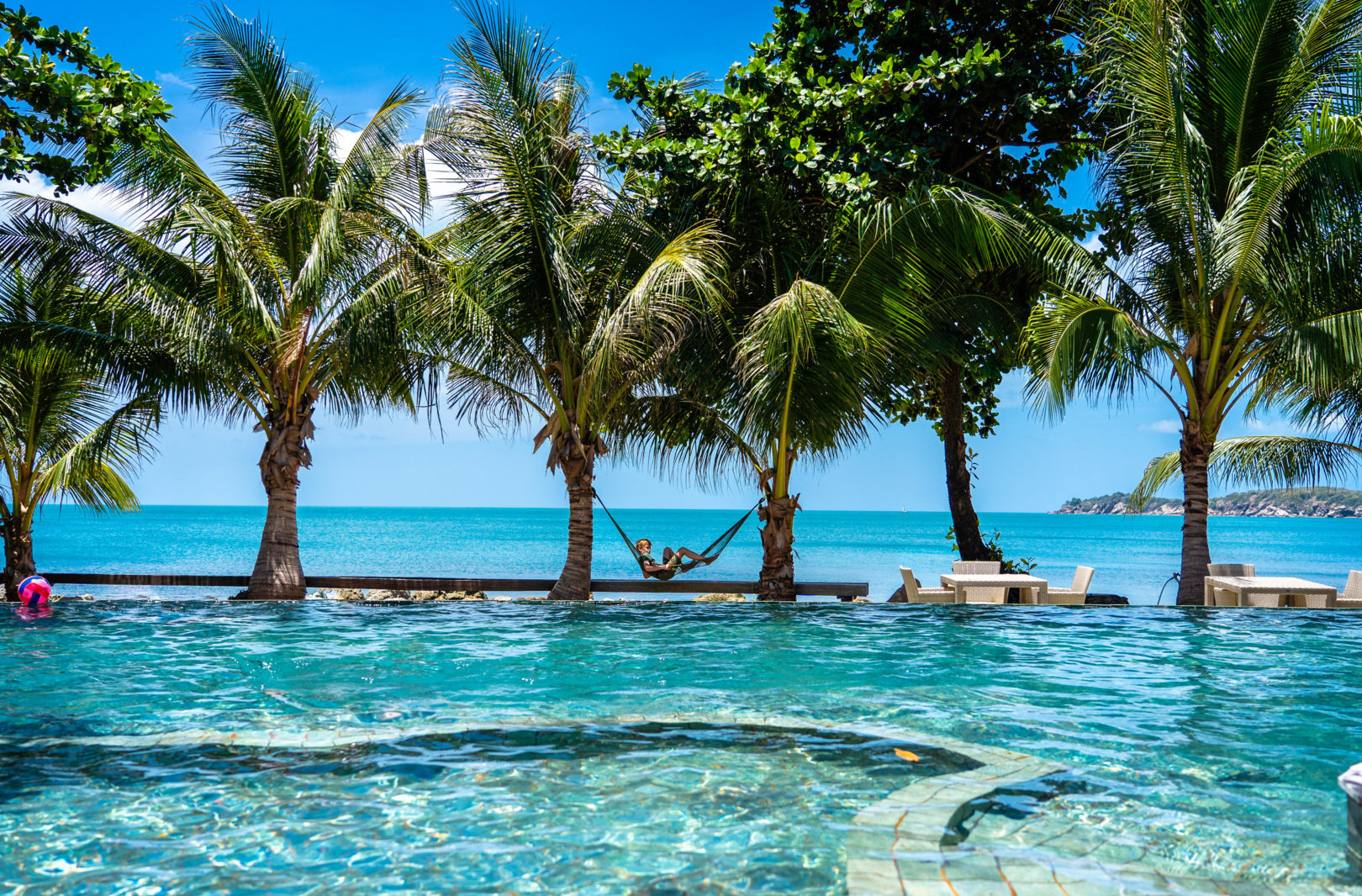 Infinity pool overlooking the ocean with palm trees lining the edge, a hammock hanging between palms, and outdoor dining tables nearby