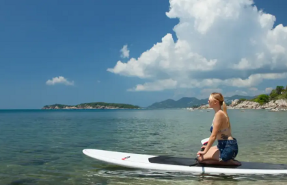 Guest enjoying calm sea on a paddleboard near a rocky beach with lush greenery and mountains under a bright blue sky