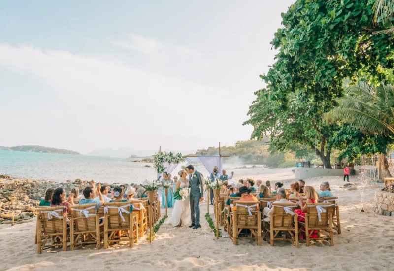 Romantic beachfront wedding ceremony on sandy beach with wooden chairs, floral arch, and guests under lush green trees