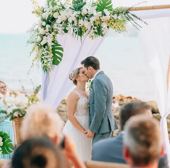 Bride and groom share a kiss under a floral arch on a beach, surrounded by guests during an elegant outdoor wedding ceremony