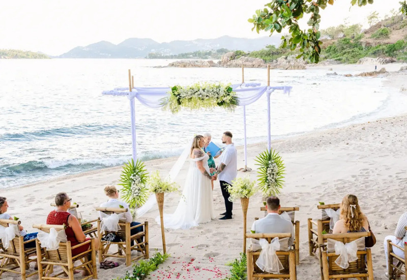 Beach wedding ceremony with couple exchanging vows under a floral arch, surrounded by bamboo chairs and ocean views