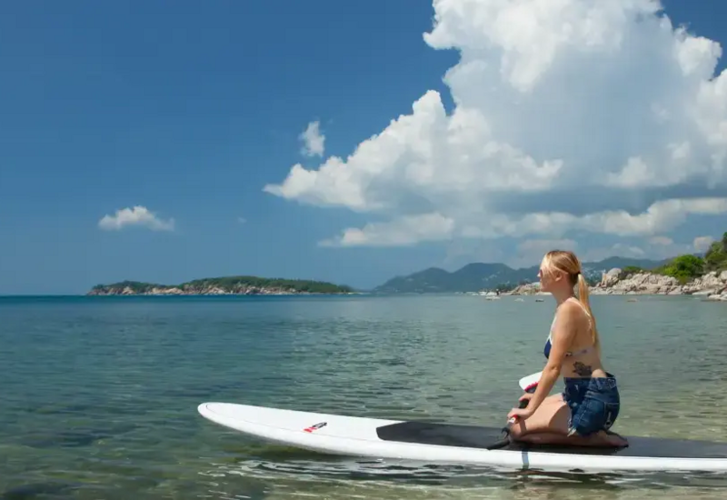 Guest enjoying calm sea on a paddleboard near a rocky beach with lush greenery and mountains under a bright blue sky
