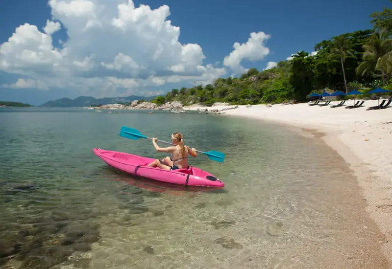 Guest kayaking in clear shallow waters along a sandy beach with lounge chairs and umbrellas under palm trees