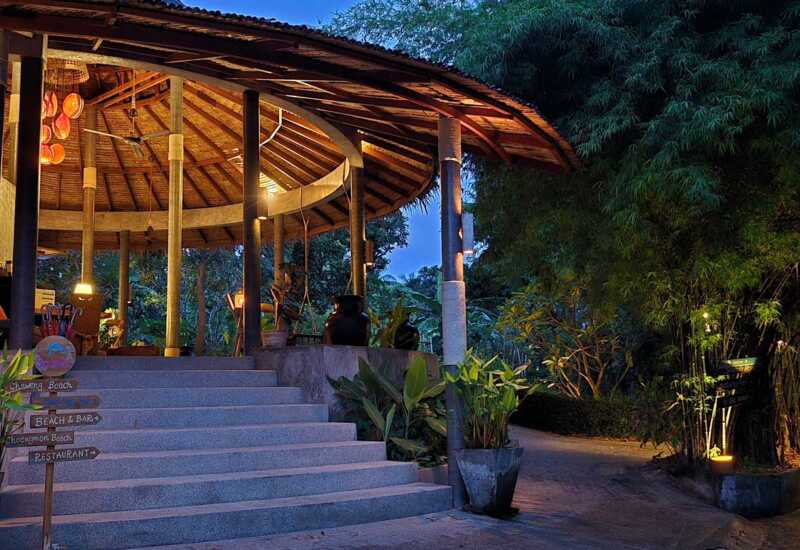 Thatched-roof open-air lounge with ambient lighting, surrounded by lush tropical plants and directional signs to beaches and restaurant