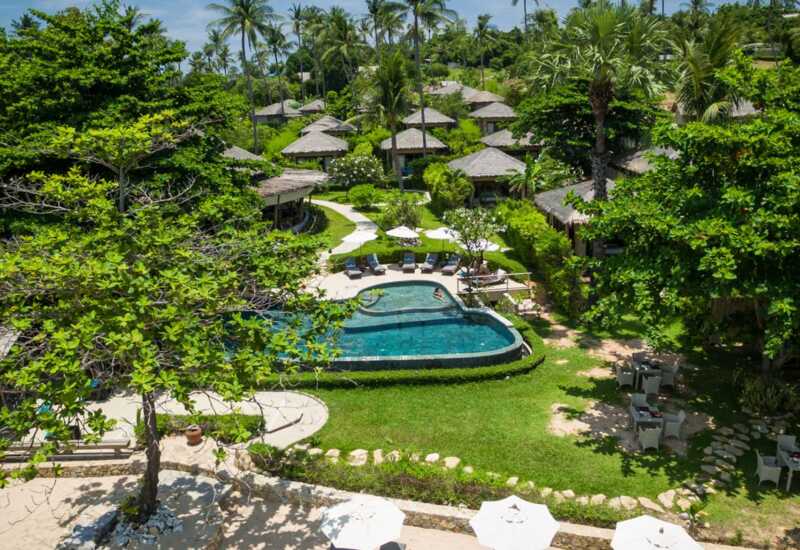 Tropical resort with private thatched-roof villas, central pool surrounded by loungers and umbrellas, lush greenery and outdoor dining areas