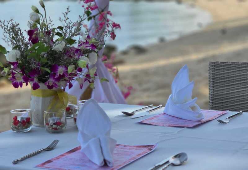 Beachfront dining table set with white linens, folded napkins, floral centerpiece, and romantic ocean view in the background
