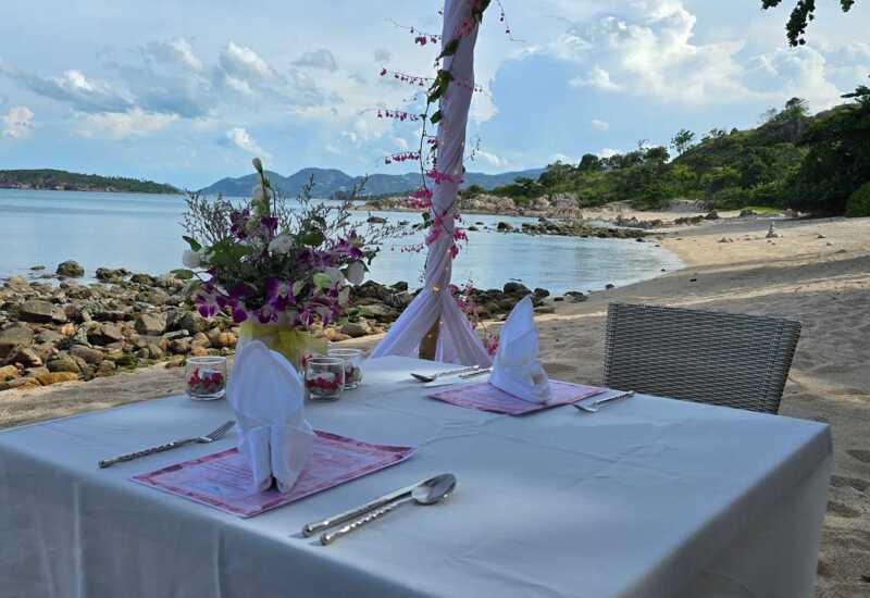 Romantic beachside dining setup with white linen table, folded napkins, floral centerpiece, and ocean views with rocky shore and hills