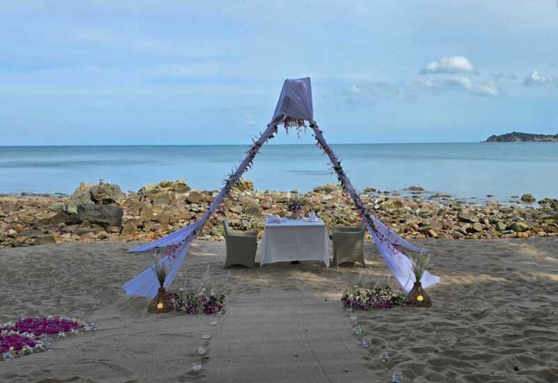 Romantic beachfront dining setup with white canopy and floral decorations on sandy shore overlooking calm sea and rocky coastline
