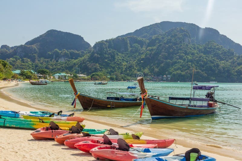 Scenic beachfront with colorful kayaks lined on sandy shore and traditional wooden boats floating on clear turquoise water near lush green hills