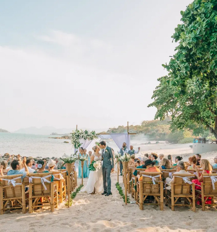 Romantic beachfront wedding ceremony on sandy beach with wooden chairs, floral arch, and guests under lush green trees
