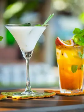 Two refreshing cocktails on a wooden table, featuring a creamy white martini and a fruity orange drink garnished with mint and passionfruit.