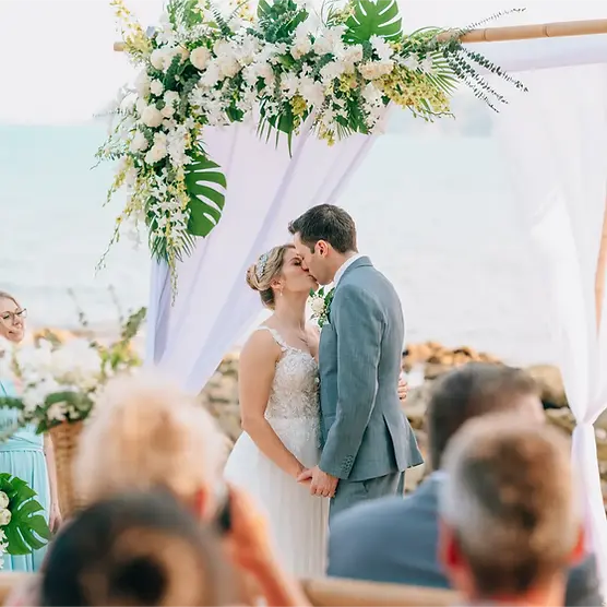 Bride and groom share a kiss under a floral arch on a beach, surrounded by guests during an elegant outdoor wedding ceremony