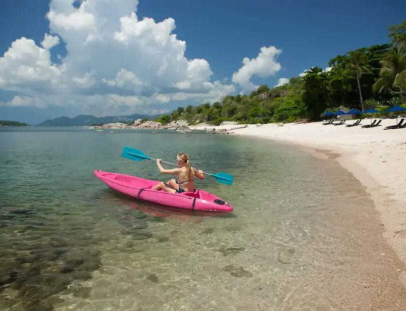 Guest kayaking in clear shallow waters along a sandy beach with lounge chairs and umbrellas under palm trees