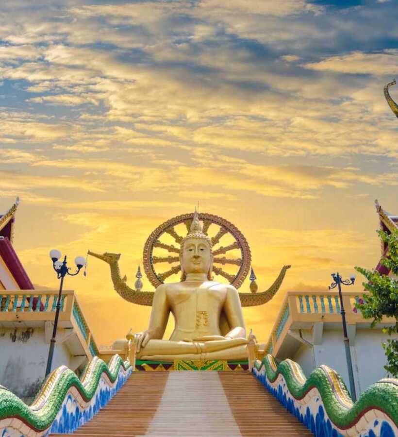 Grand golden Buddha statue seated at the top of ornate staircase with detailed serpent railings and traditional temple roofs at sunset