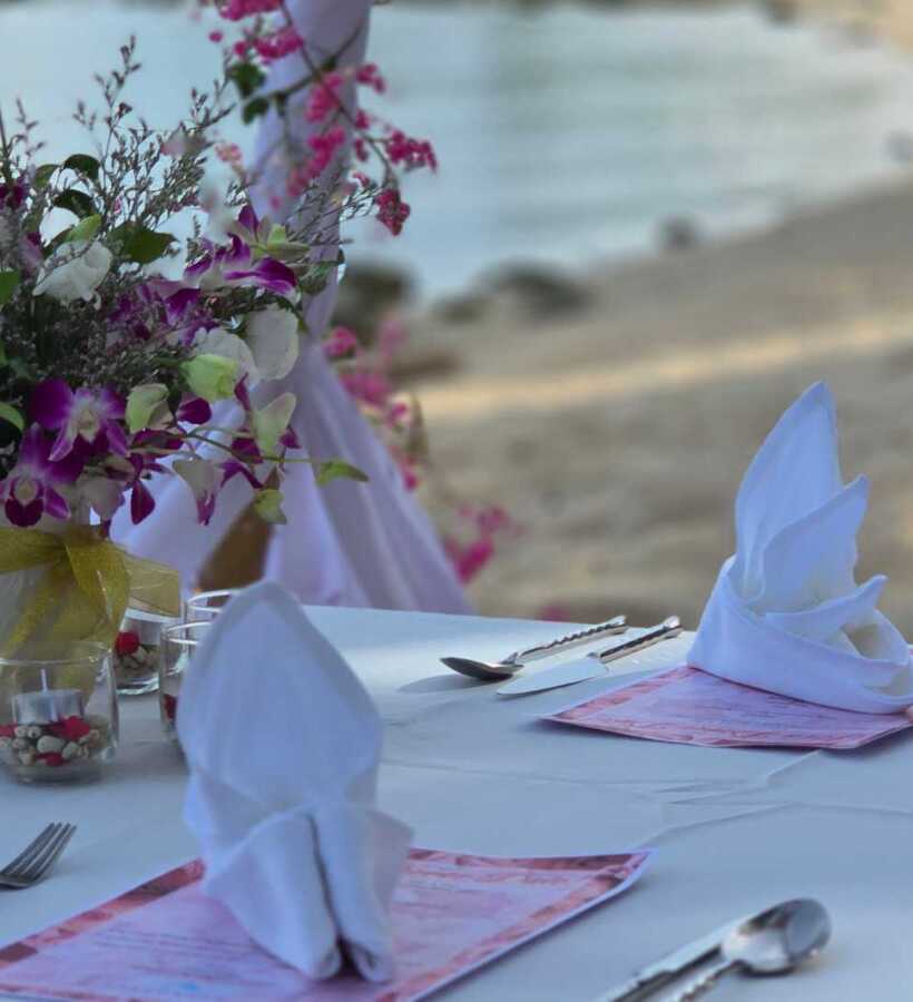 Beachfront dining table set with white linens, folded napkins, floral centerpiece, and romantic ocean view in the background