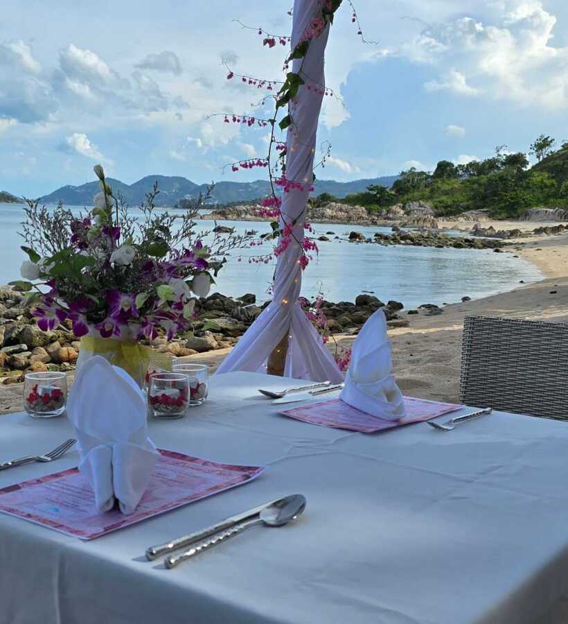 Romantic beachside dining setup with white linen table, folded napkins, floral centerpiece, and ocean views with rocky shore and hills