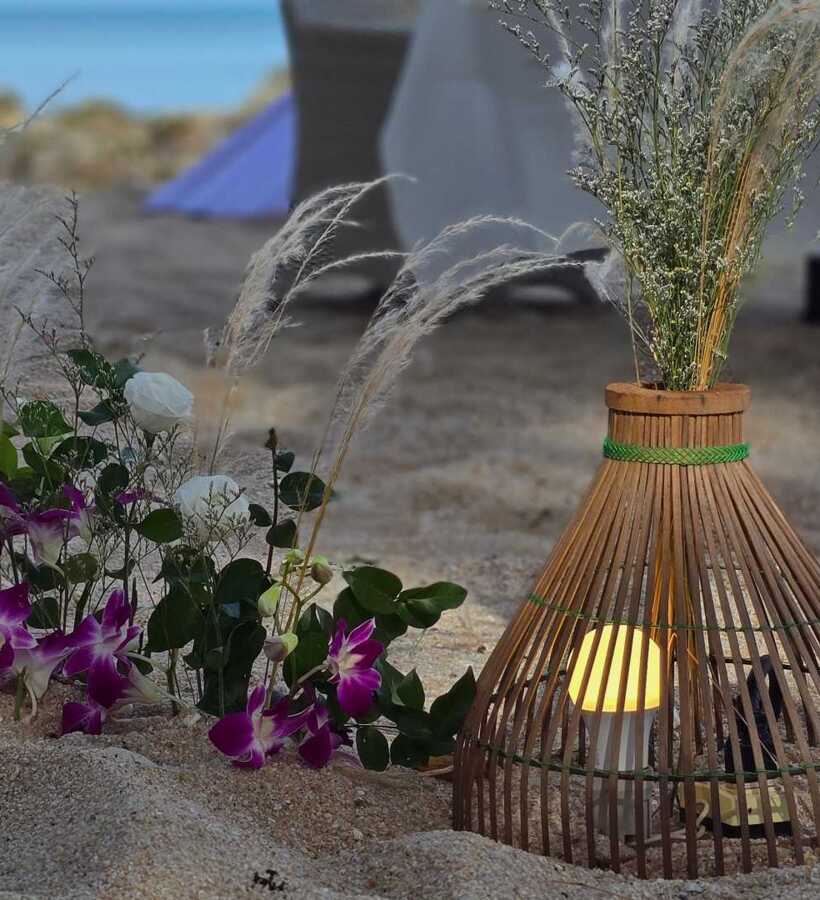 Beachside floral arrangement with purple and white flowers next to a glowing lantern and dried grasses in sand, setting a romantic ambiance