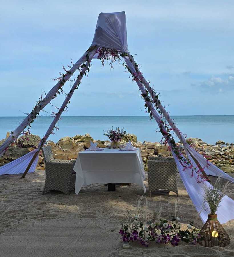 Romantic beachside dining setup with white tablecloth, floral centerpiece, wicker chairs, and draped canopy by calm sea and rocky shore
