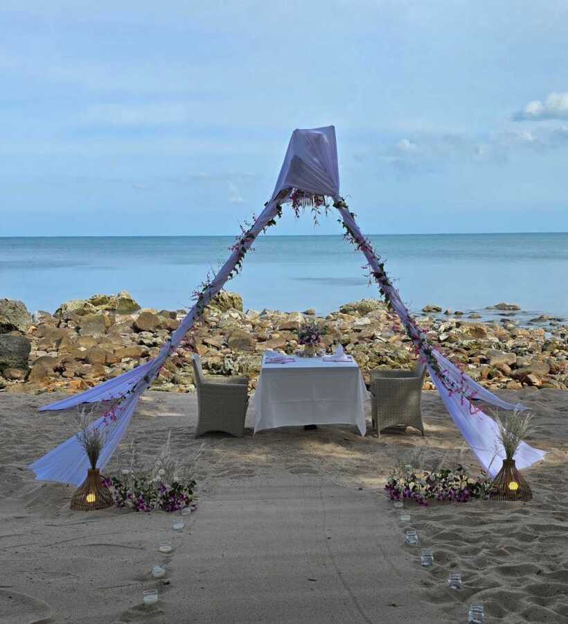 Romantic beachfront dining setup with white canopy and floral decorations on sandy shore overlooking calm sea and rocky coastline