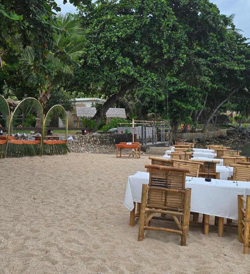 Beachfront dining setup with bamboo chairs and white tablecloths on sand, surrounded by trees and ocean view