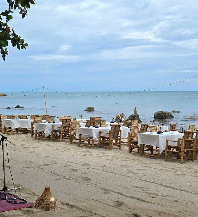 Beachfront dining with wooden tables and chairs set for multiple guests, string lights overhead, calm sea and rocky shoreline in background