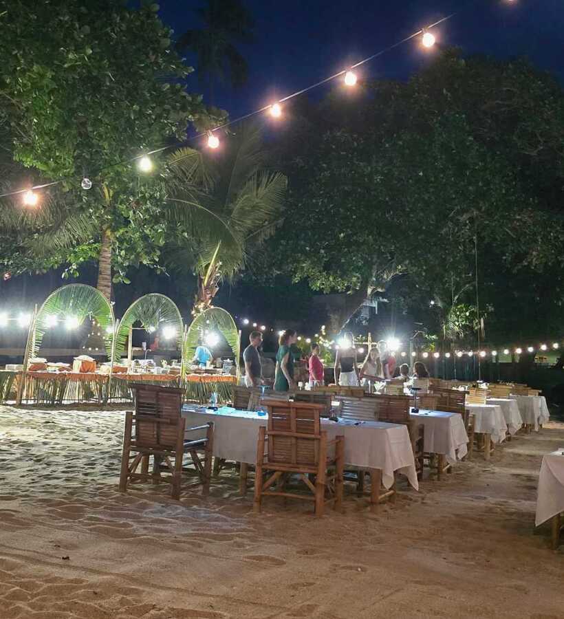 Beachside dining setup on sand with wooden chairs and tables under string lights, guests enjoying a relaxed evening meal