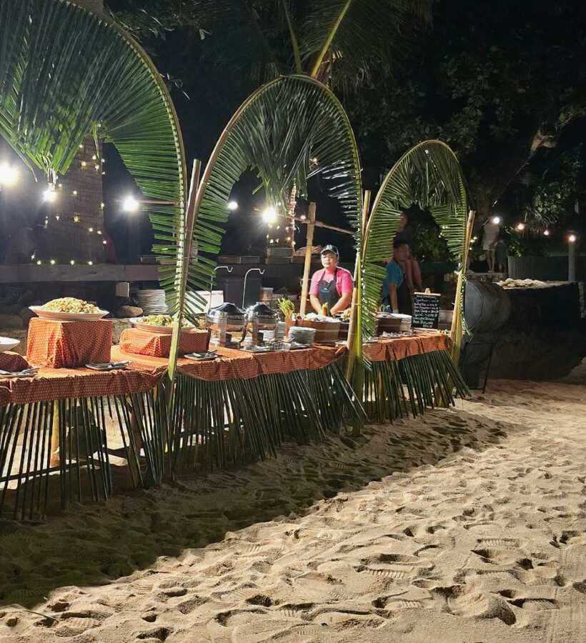 Outdoor nighttime beach buffet with tropical palm leaf arches, warm lighting, and staff preparing food on sand under string lights