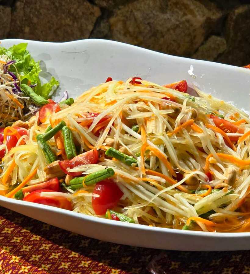 Fresh Thai papaya salad with cherry tomatoes, long beans, shredded carrots, and mixed greens on a white dish