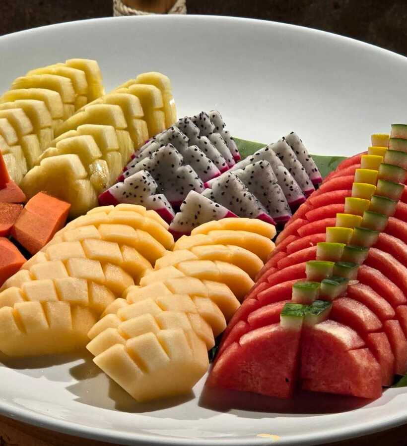 Platter of fresh tropical fruits including papaya, pineapple, dragon fruit, melon, and watermelon, arranged on banana leaf on white plate