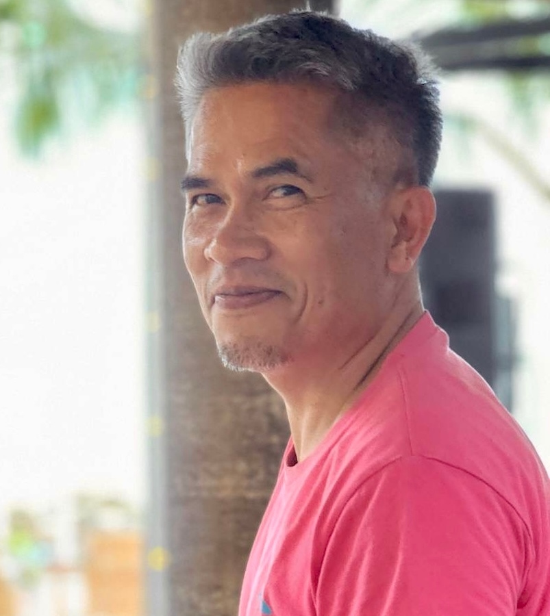 Man wearing a pink shirt smiling with a blurred background, possibly at a tropical or outdoor hotel setting