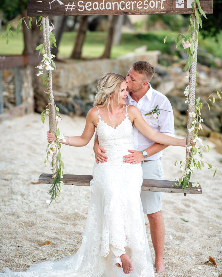 Newlywed couple in wedding attire on a floral-decorated beach swing at Sea Dance Resort, sharing a romantic moment by the shore