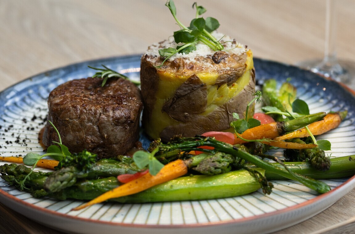 Gourmet fillet steak with layered potato terrine, asparagus, baby carrots and microgreens on a ceramic plate, wine glass at edge.