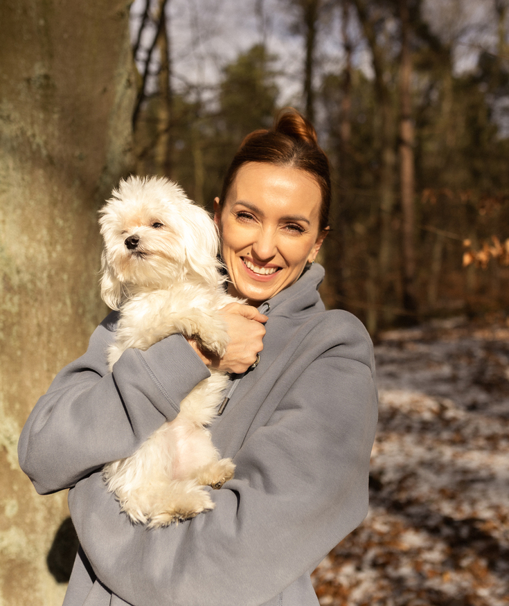 Smiling woman in a hoodie holding a small white dog during a sunlit winter walk in a forest