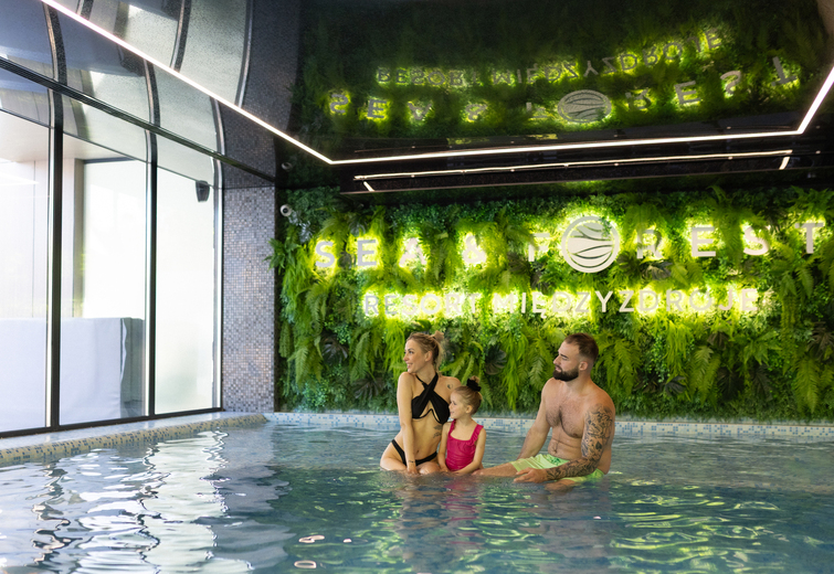 Family relaxing in a hotel indoor pool beside an illuminated living green wall with 'Sea & Forest' sign, spa ambiance