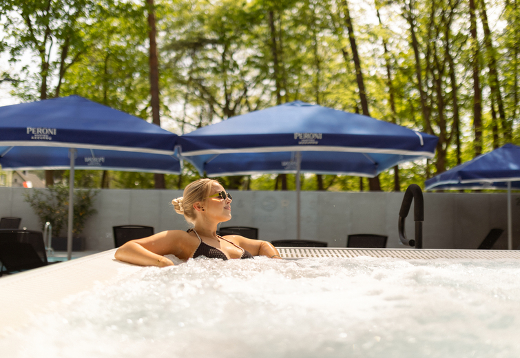 Woman relaxing in bubbling outdoor jacuzzi by blue pool umbrellas, lounge chairs and leafy trees.