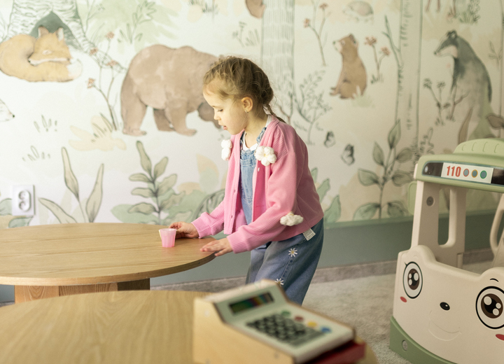 Children's playroom with forest wall mural, wooden tables, toy cash register and a girl playing with a pink cup