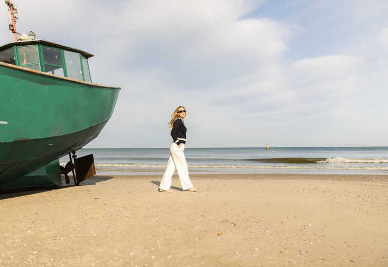 Woman in sunglasses strolling on a wide sandy beach beside a large green beached fishing boat, calm sea and clear sky