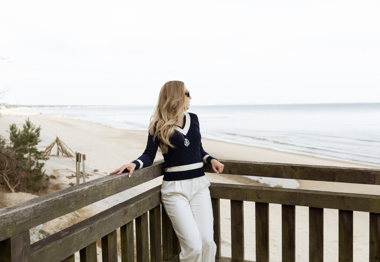 Woman in sunglasses leaning on a wooden balcony overlooking a calm sandy beach and sea, relaxed beachfront terrace vibe