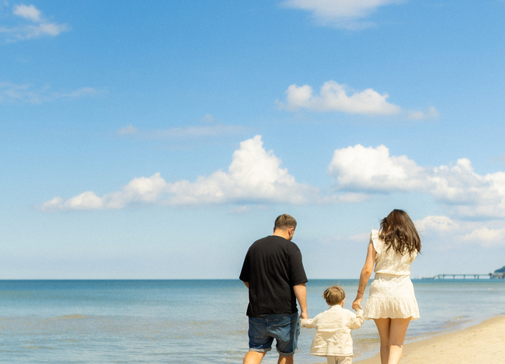 Parents and child walking barefoot on a sandy hotel beach, calm sea, blue sky, relaxed family-friendly atmosphere