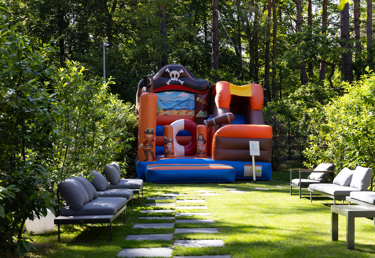 Sunlit hotel garden with stepping-stone lawn, lounge seating and a colorful pirate-themed inflatable bouncy castle for children.