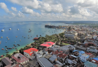 Coastal cityscape with historic buildings, red roofs, boats in calm blue harbor, and green hills in the distance under a partly cloudy sky