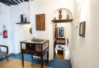 Bright hallway with white walls, wooden decorative shelves, glass display cabinet, colorful pillow on gray chair, and rustic ceiling beams