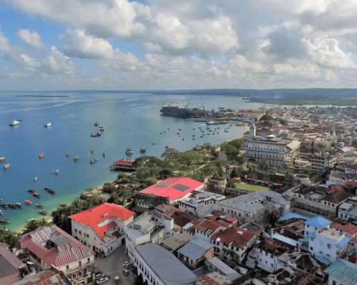 Coastal cityscape with historic buildings, red roofs, boats in calm blue harbor, and green hills in the distance under a partly cloudy sky